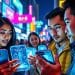 Grupo de jóvenes usando smartphones transparentes con hologramas en una calle iluminada con letreros de neón – Mundo Chido