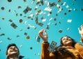 Pareja joven celebrando mientras llueven billetes de dólar y monedas sobre un fondo azul brillante – Mundo Chido