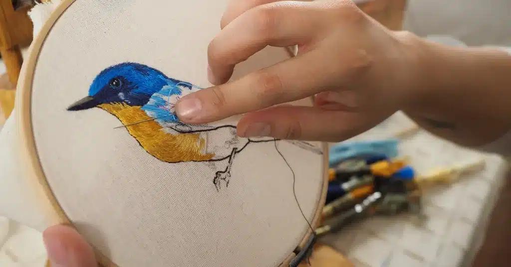 Mano bordando pájaro en bastidor – Mundo Chido - Planeta Cortes Primer plano de una mano bordando un pájaro azul y amarillo sobre un bastidor de tela.