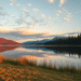 La luz dorada del sol ilumina la hierba junto a un tranquilo lago, con pinos y montañas lejanas reflejados en el agua bajo un cielo salpicado de nubes al amanecer o al atardecer.