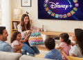Una familia de cinco miembros se sienta en un sofá, ríen y comparten palomitas mientras ven juntos Disney+ en un acogedor salón. Una mujer sostiene una manta de colores y los niños sonríen entusiasmados.