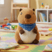 Un capibara de peluche con un pañuelo azul está sentado sobre una alfombra de espuma de colores en una habitación de juegos luminosa y ordenada, con libros, bloques de madera y estanterías de juguetes al fondo.