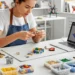 Una persona con delantal monta coloridos ladrillos de LEGO en un escritorio, rodeado de organizados contenedores de piezas de LEGO. Cerca, en un espacio de trabajo moderno y luminoso, hay un ordenador portátil con un modelo en 3D.