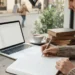 Una persona está sentada en la mesa de un café junto a una ventana, escribiendo en un cuaderno. Sobre la mesa hay un portátil abierto, libros apilados, una taza de café y un smartphone. Fuera, la gente camina por una calle adoquinada.