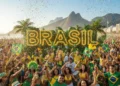 Una animada multitud celebra en Brasil, vestida de amarillo y verde, ondeando banderas y rodeada de confeti. Grandes letras BRASIL iluminadas y palmeras se alzan frente a las montañas en un día soleado.