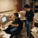 Dos jóvenes trabajan en una oficina desordenada llena de viejos monitores de ordenador, papeles y latas de refresco. Uno teclea en un escritorio que muestra una página de búsqueda de Google, mientras el otro permanece de pie a su lado, gesticulando animadamente.