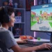 Un niño sonriente con auriculares está sentado en un escritorio, jugando al colorido juego Adopt Me! en un gran monitor. Sobre el escritorio, en una acogedora habitación decorada, hay aperitivos, un teclado y un ratón.